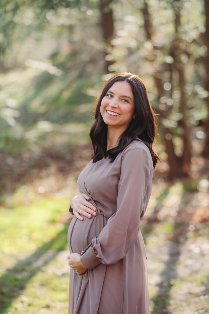 Pregnant woman in a flowing dress holding her baby bump, standing in soft golden light around 30 weeks of pregnancy