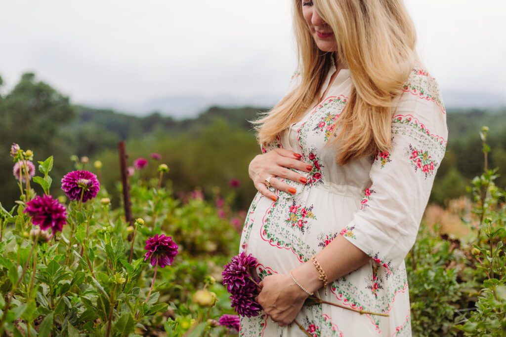 Pregnant woman in a flower field holding her baby bump, standing in soft golden light around 32 weeks of pregnancy