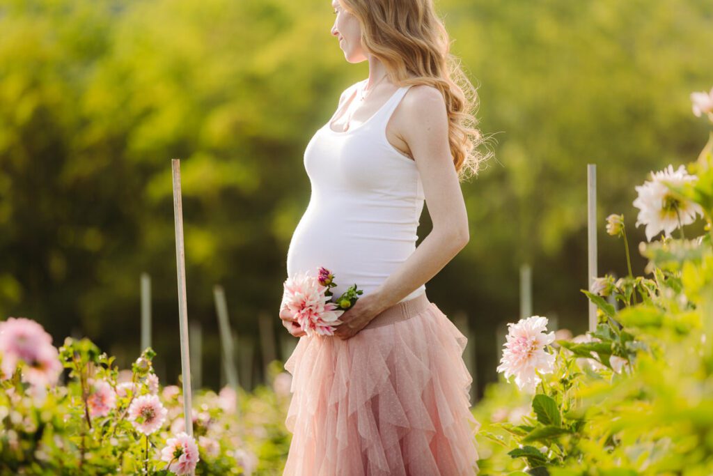 Pregnant woman in a flower field holding her baby bump, standing in soft golden light around 30 weeks of pregnancy