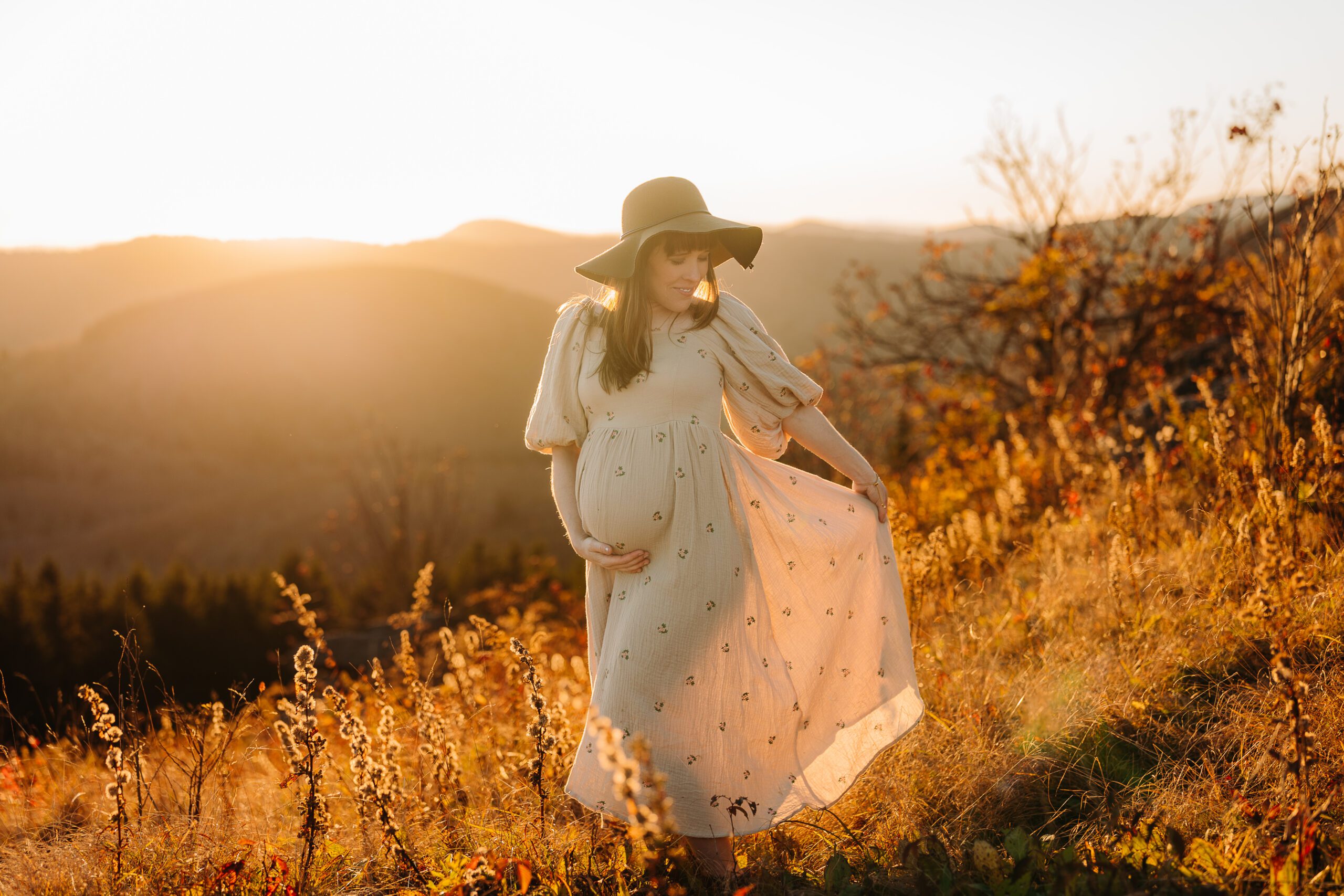 Beautiful maternity photo session with woman in beige floral dress and floppy hat on a mountaintop