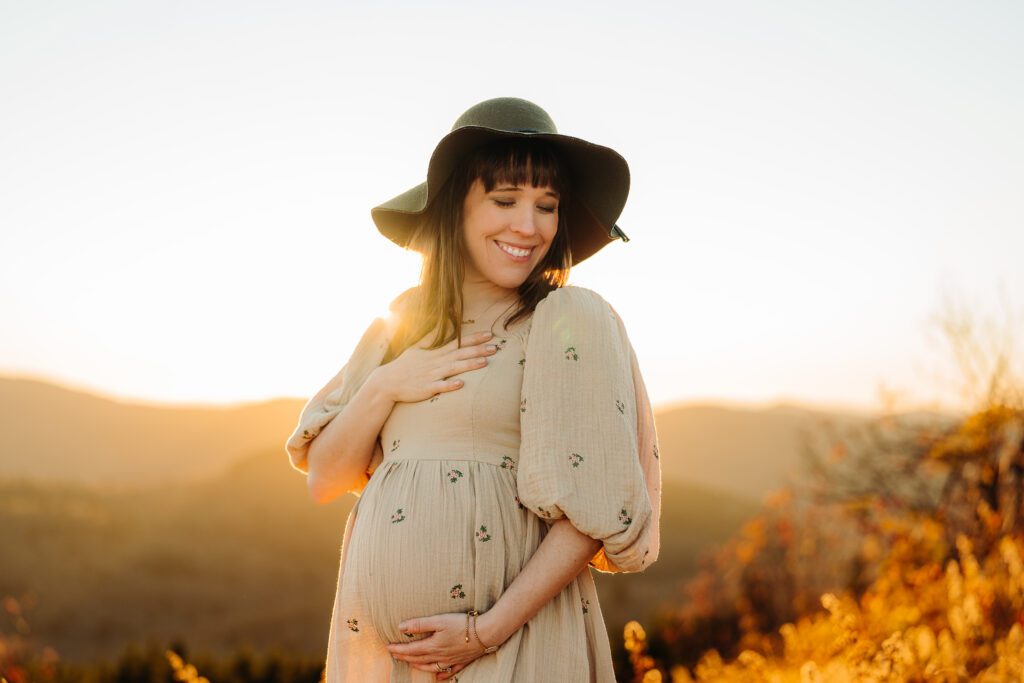 Pregnant woman in beige dress and floppy hat on a mountaintop at sunset.